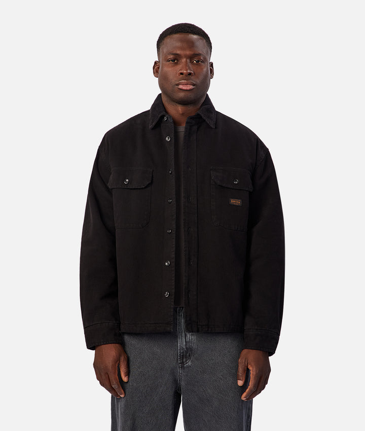 A man wearing a black button-up overshirt jacket with flap chest pockets and gray jeans, standing against a light gray studio background.